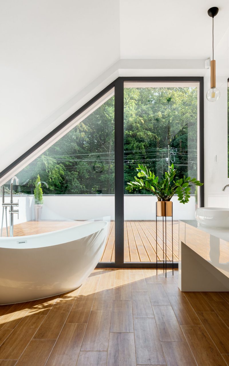Elegant attic bathroom with stylish bathtub, wooden floor and balcony door